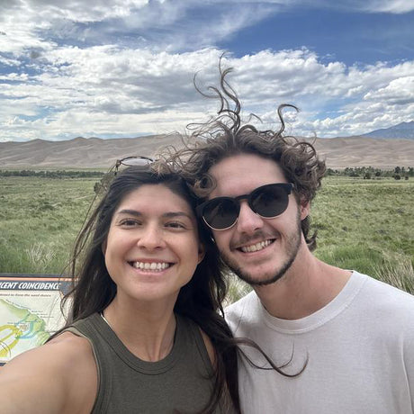 Two people posing for a selfie with a scenic background of mountains and open fields.