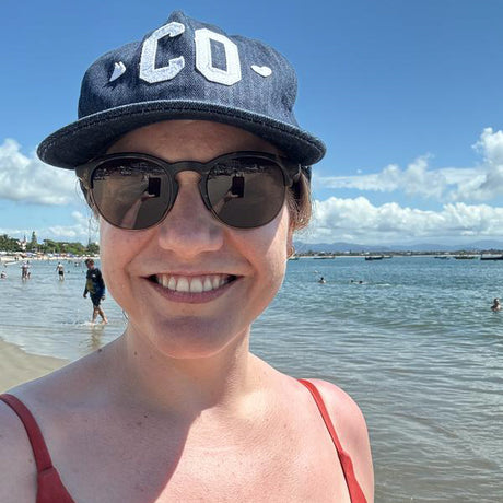 Person wearing a cap and sunglasses at a beach with water and sky in the background