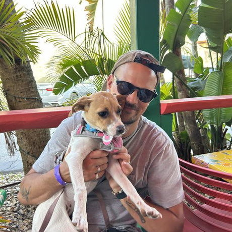 Man holding a small dog outdoors with greenery in the background
