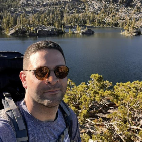 Man with sunglasses and backpack standing in front of a lake and forest
