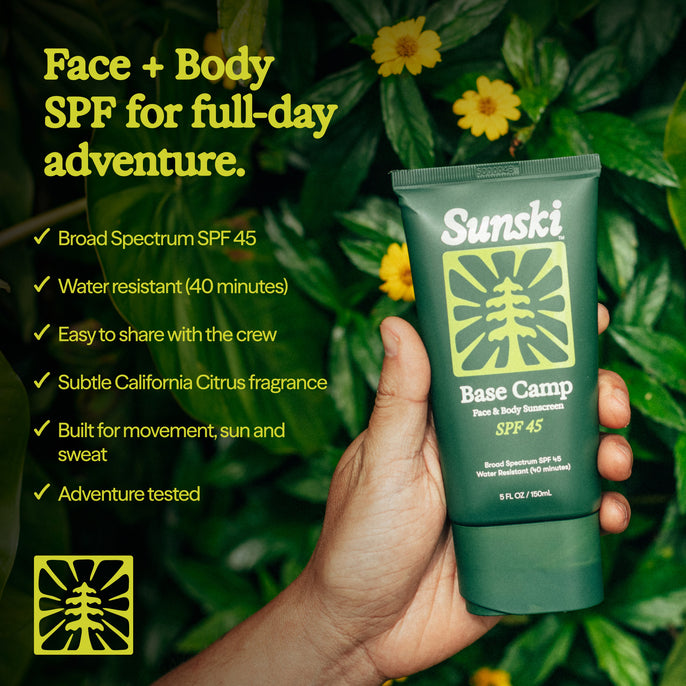 A hand holds a green Sunski Base Camp Face + Body SPF 45 tube against a background of green foliage with yellow flowers. Yellow text on the left lists product benefits.