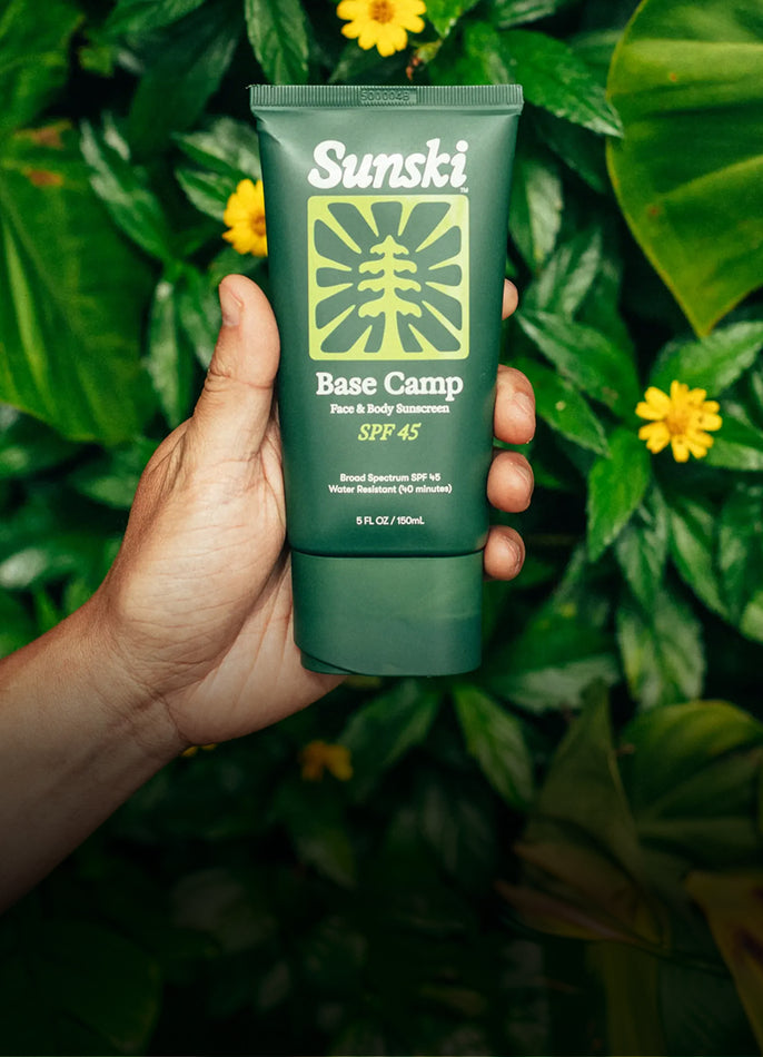 A hand holding a green tube of Sunski Base Camp SPF 45 sunscreen against a background of lush green tropical leaves and small yellow flowers.