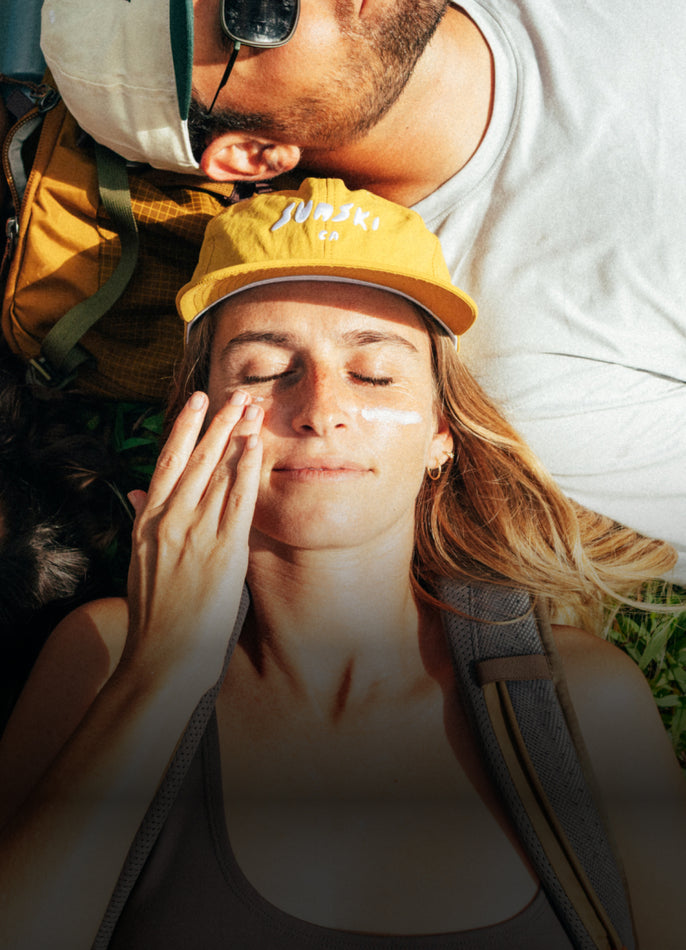 A woman lying outdoors in the sun wearing a yellow baseball cap and applying sunscreen with her hand
