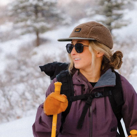 Person hiking in a snowy landscape wearing a brown cap, sunglasses, and purple jacket.