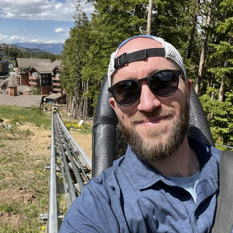 Man with sunglasses and a beanie standing outdoors with trees and buildings in the background