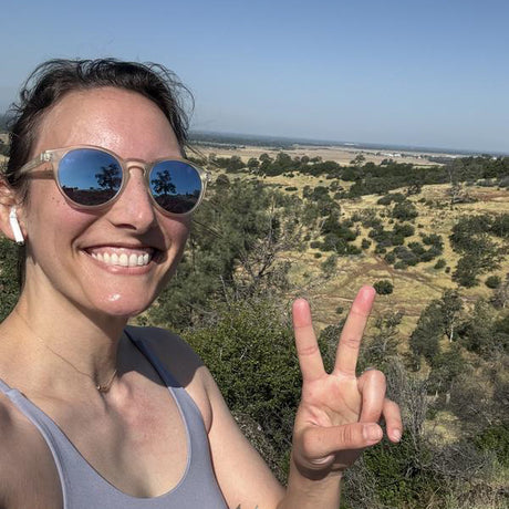 Woman wearing sunglasses and a gray tank top, making a peace sign with her hand, with a scenic landscape in the background.