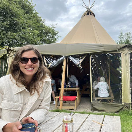 Woman sitting at a table with a cup, smiling in front of a tipi tent.