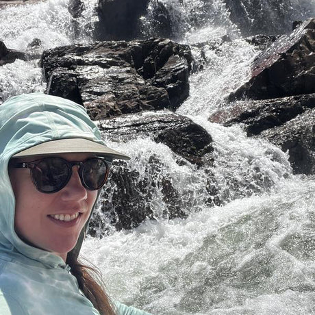 Person wearing a light blue hooded jacket and sunglasses in front of a flowing river with rocks.