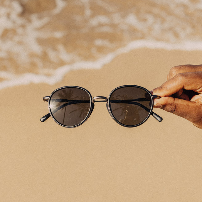Sunglasses held by a hand on a sandy beach with waves in the background