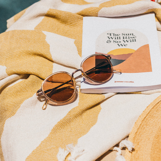 Sunglasses on a book titled 'The Sun Will Rise & So Will We' on a yellow and white patterned fabric.