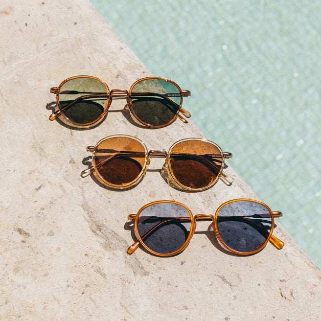 Three pairs of sunglasses with gold frames on a stone surface near water.
