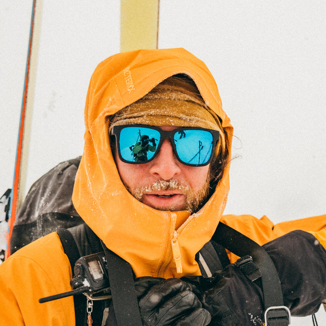 Person wearing an orange hooded jacket and blue sunglasses with a snowy background