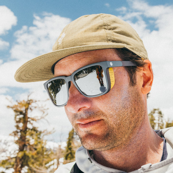 Man wearing sunglasses and a cap outdoors with trees and sky in the background