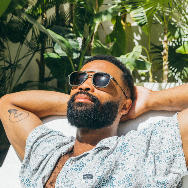 Man wearing sunglasses and a patterned shirt, lying on a couch with plants in the background