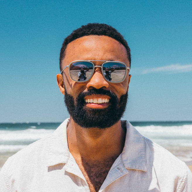 Man wearing sunglasses and a light shirt on a beach with clear blue sky and ocean.