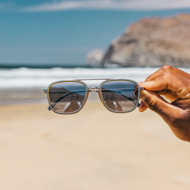 Sunglasses held by a hand with a beach and ocean in the background