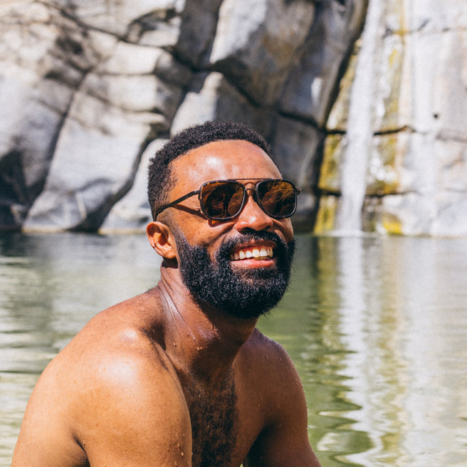 Man wearing sunglasses by a body of water with rocky cliffs in the background