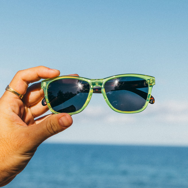 Hand holding green sunglasses with a clear blue sky and ocean background