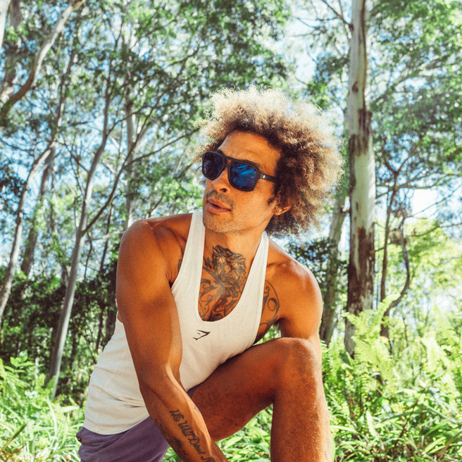Man with tattoos wearing a white tank top and sunglasses in a forest setting