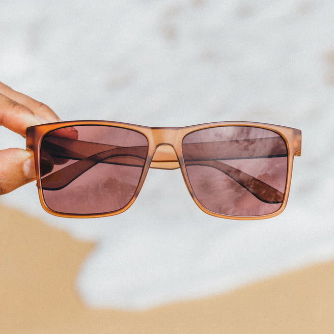 Sunglasses with brown frame and pink-tinted lenses held over a beach scene.