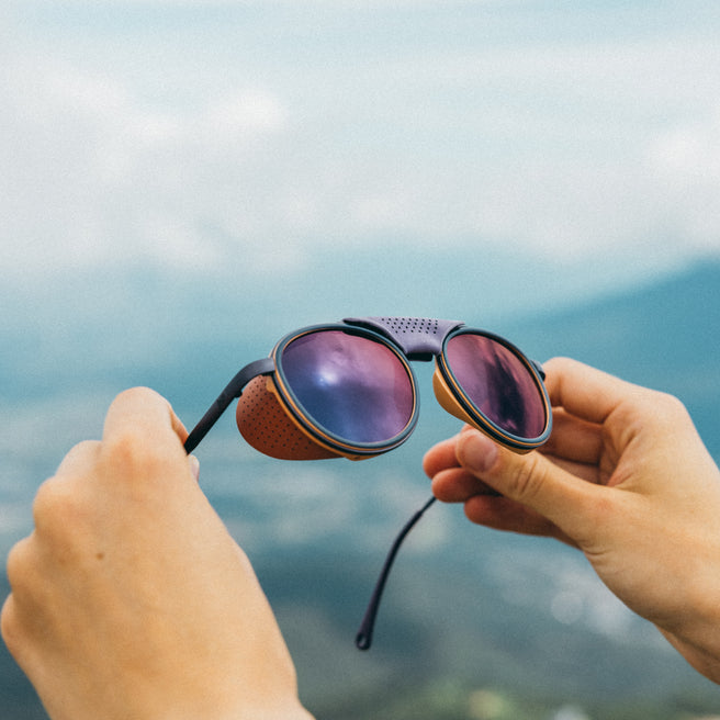 Sunglasses with purple lenses held by hands against a blurred natural background
