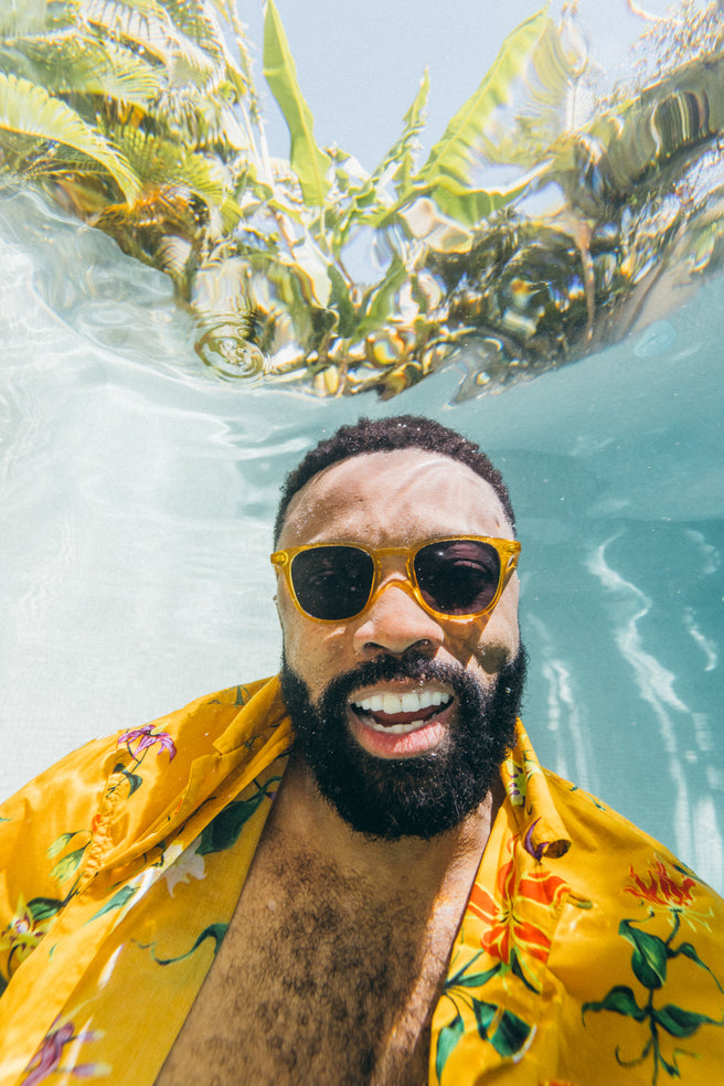 Man wearing sunglasses and a yellow floral shirt underwater with seaweed.