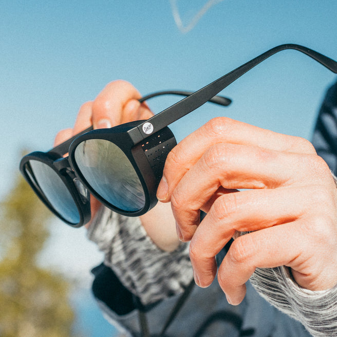 Person holding a pair of black sunglasses with a blurred outdoor background