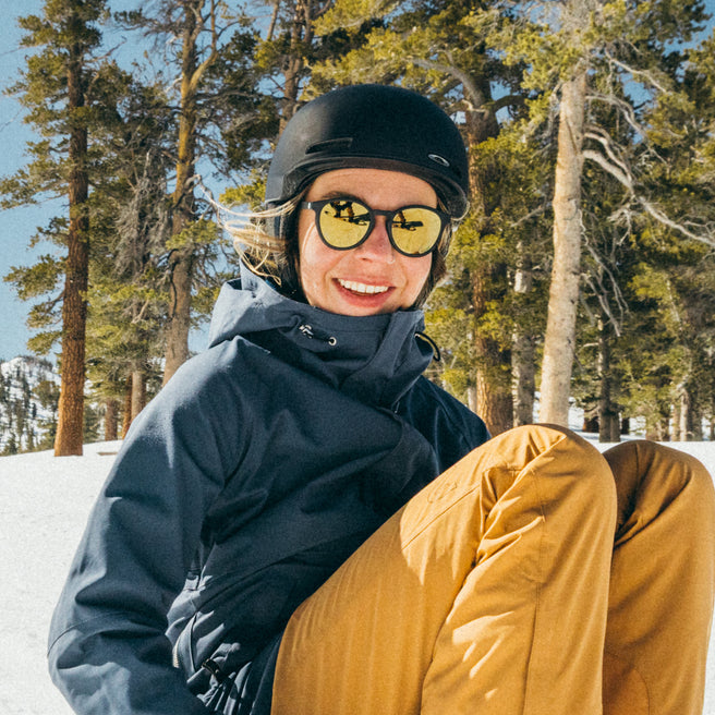 Person in winter gear with yellow sunglasses sitting on a snowy ground with trees in the background