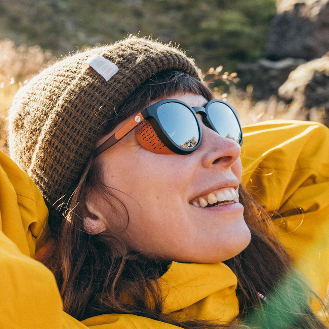 Person wearing a brown beanie and yellow jacket with sunglasses, outdoors.