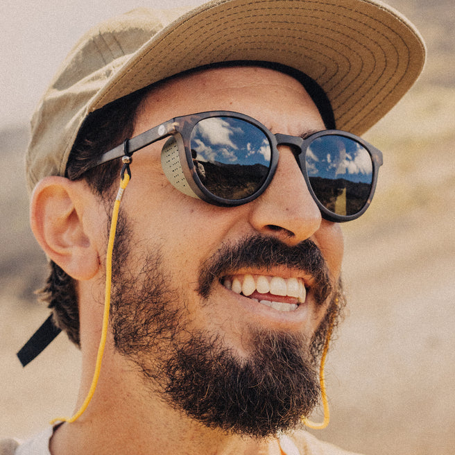 Man wearing sunglasses with a reflection of clouds and mountains, wearing a cap and smiling.