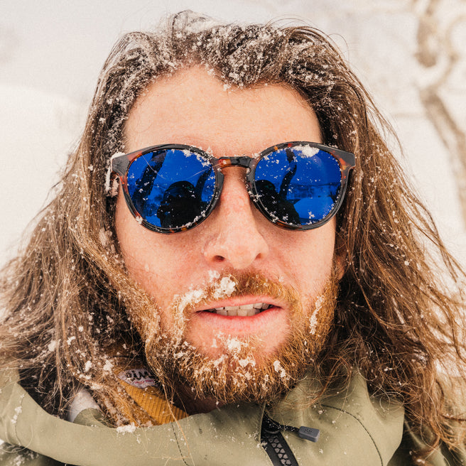 Person with snow-covered hair and beard wearing blue sunglasses in a snowy environment