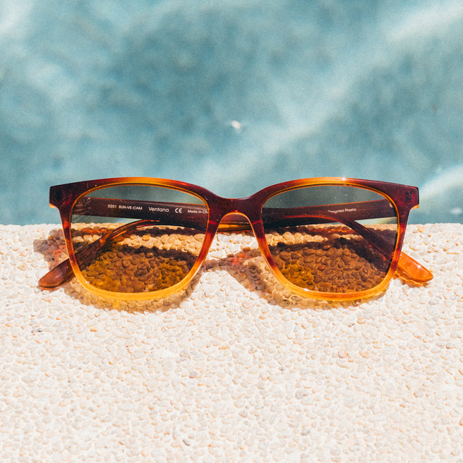 Sunglasses with a tortoiseshell frame on a sandy surface with a blurred blue background