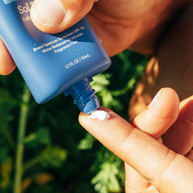 Sol Power SPF 50 Sunscreen - 1.7 Fl oz / 50 mL - Person applying sunscreen to their hand with a blue bottle against a blurred green background