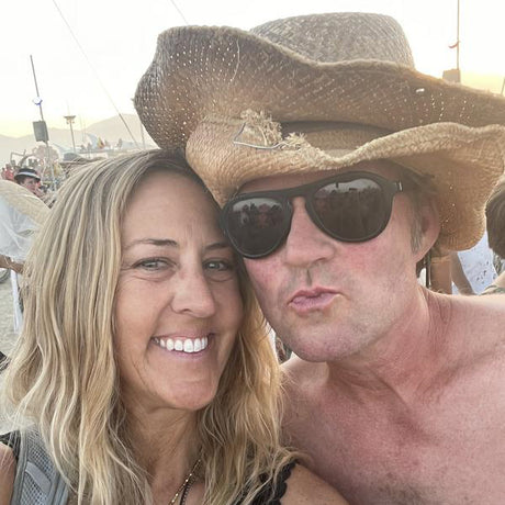 Two people posing for a selfie, one wearing a cowboy hat and sunglasses, with a blurred background.