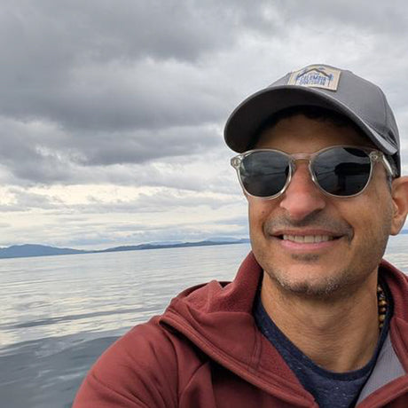 Man wearing sunglasses and a cap with a scenic background of water and mountains.