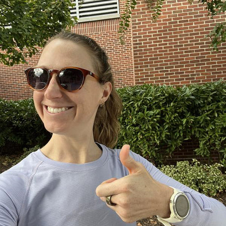 Person wearing sunglasses and a light purple shirt, giving a thumbs-up in front of a brick wall and greenery.
