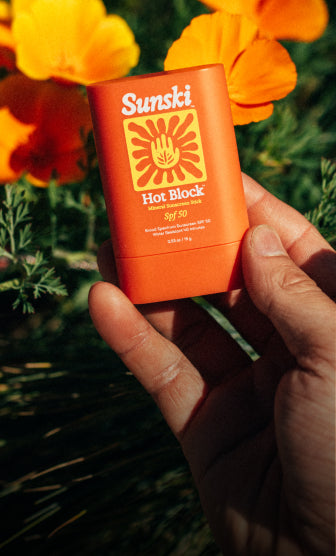 Hand holding a Sunski Hot Block sunscreen stick with flowers in the background