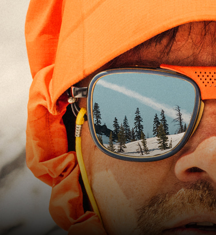 Person wearing orange goggles with a reflection of trees and sky, likely in a snowy landscape.